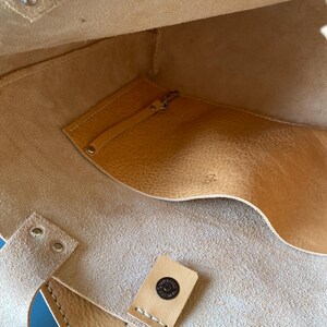 May include: The inside of a light brown leather tote bag with a small pocket and a snap closure. The bag has a stitched seam and a leather tag with the text "Seattle Leather Co."