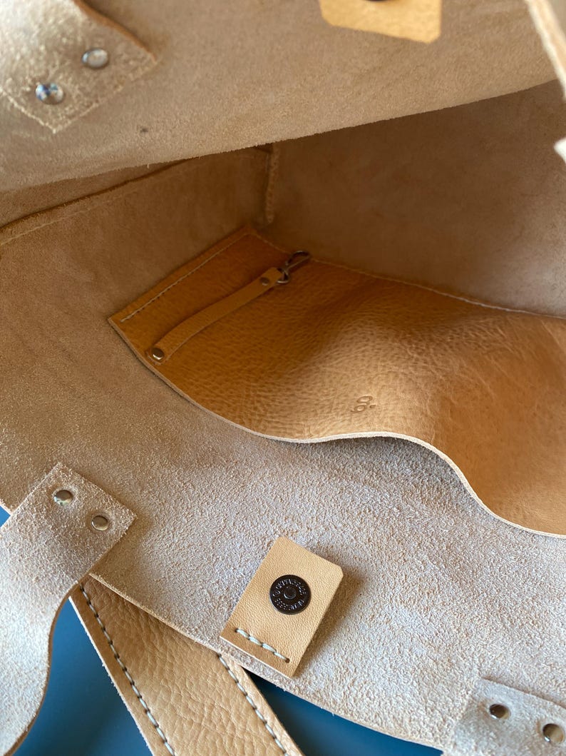 May include: The inside of a light brown leather tote bag with a small pocket and a snap closure. The bag has a stitched seam and a leather tag with the text "Seattle Leather Co."