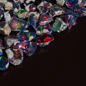 Op de afbeelding: Een verzameling zwarte opaal edelstenen met iriserende regenboogkleuren en rode flitsen. De opalen zijn ruw en hebben een natuurlijke, onbewerkte uitstraling.