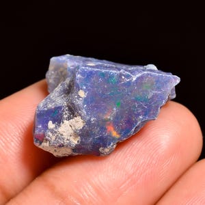 Könnte beinhalten: Nahaufnahme eines rauen, ungeschliffenen Opal-Edelsteins. Der Opal zeigt eine dunkelblaue Grundfarbe mit grünen, orangefarbenen und roten Farbblitzen. Der Stein hat ein rohes, natürliches Aussehen mit einer strukturierten Oberfläche.