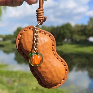 Könnte beinhalten: Handgefertigter Lederanhänger in Erdnussform mit einem kleinen orangefarbenen Glasanhänger. Der Anhänger ist an einer braunen Lederschnur befestigt. Im Hintergrund sind ein Fluss und ein blauer Himmel zu sehen.