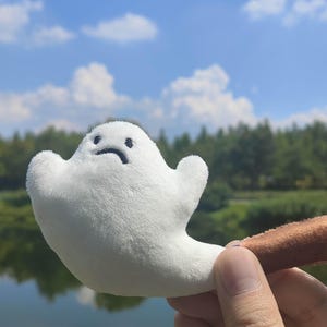Op de afbeelding: Een witte pluche geest met een verdrietige uitdrukking, bevestigd aan een bruine cilindrische sleutelhanger. De geest wordt omhoog gehouden tegen een achtergrond van een blauwe lucht, bomen en een watermassa.
