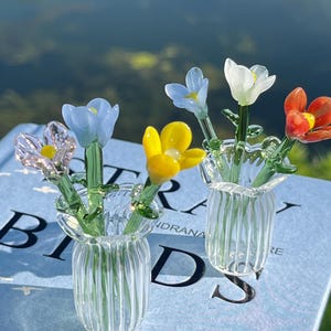 May include: Two small glass vases, each holding colorful glass flowers. The flowers include blue, yellow, white, and red blooms. The vases are clear with a ribbed design. The background shows a body of water and greenery.