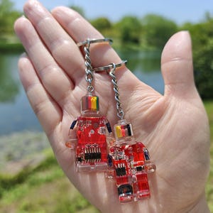 May include: Two keychains featuring robot-shaped charms. The charms are red and transparent, with internal circuit board designs. Each robot has a colorful top and is attached to a silver chain and keyring. The background is a blurred outdoor scene.