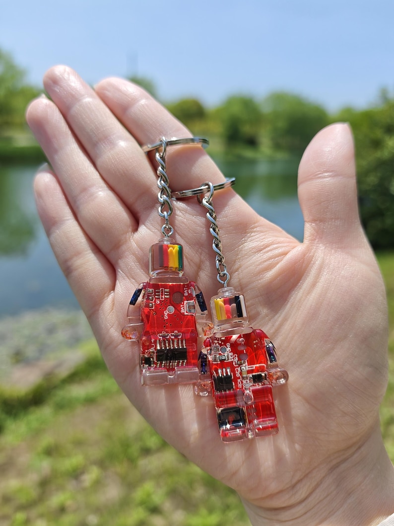 May include: Two keychains featuring robot-shaped charms. The charms are red and transparent, with internal circuit board designs. Each robot has a colorful top and is attached to a silver chain and keyring. The background is a blurred outdoor scene.