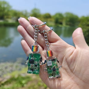 May include: Two robot-shaped keychains. Each keychain features a clear body with a green circuit board design and a rainbow-colored top. The keychains are attached to silver chains and key rings. The background shows a body of water and trees.
