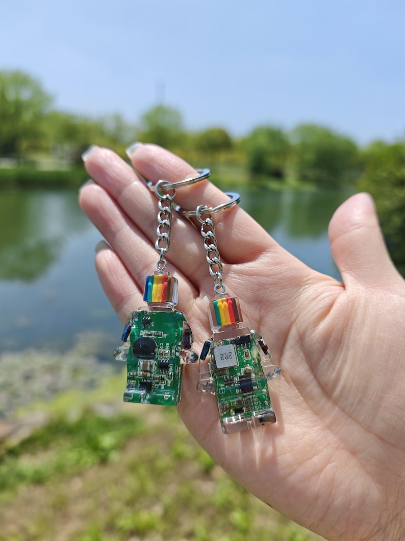 May include: Two robot-shaped keychains. Each keychain features a clear body with a green circuit board design and a rainbow-colored top. The keychains are attached to silver chains and key rings. The background shows a body of water and trees.