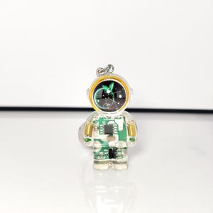 May include: A clear resin keychain shaped like an astronaut, featuring an embedded green circuit board pattern throughout the body. The helmet visor is black with a white constellation design, accented by a gold-colored rim and small metal springs.
