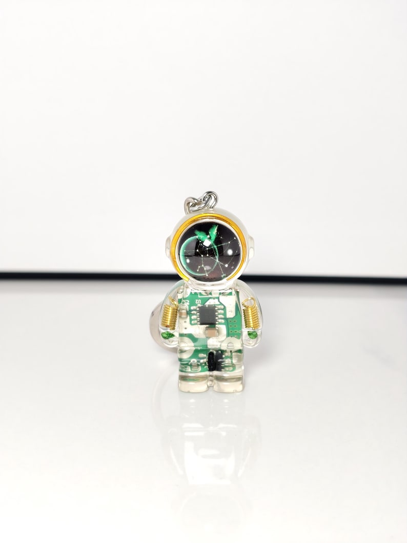May include: A clear resin keychain shaped like an astronaut, featuring an embedded green circuit board pattern throughout the body. The helmet visor is black with a white constellation design, accented by a gold-colored rim and small metal springs.
