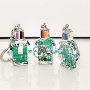 May include: Three robot-shaped keychains. Each keychain has a clear body with a green circuit board design inside. The robot heads are silver with a rainbow stripe. The arms are small, light blue crystals. The keychains are on a silver chain.