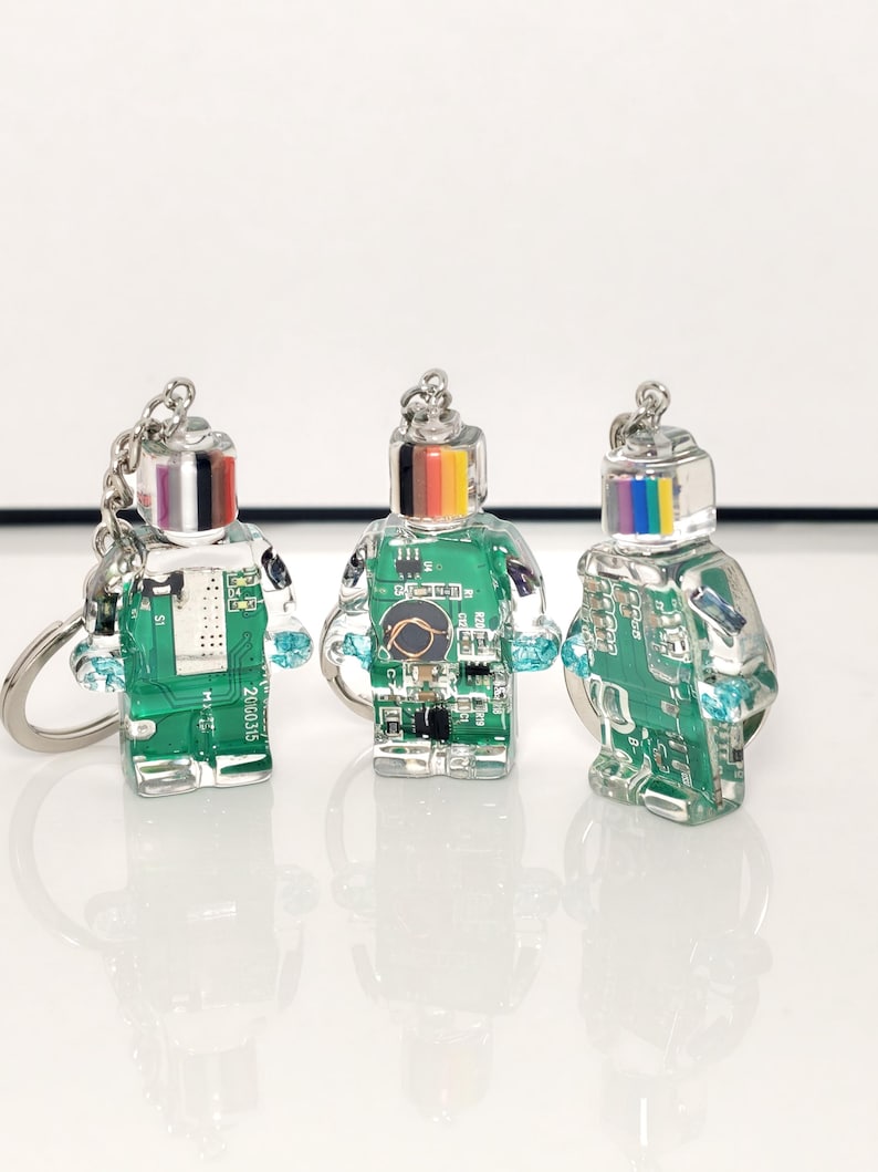 May include: Three robot-shaped keychains. Each keychain has a clear body with a green circuit board design inside. The robot heads are silver with a rainbow stripe. The arms are small, light blue crystals. The keychains are on a silver chain.
