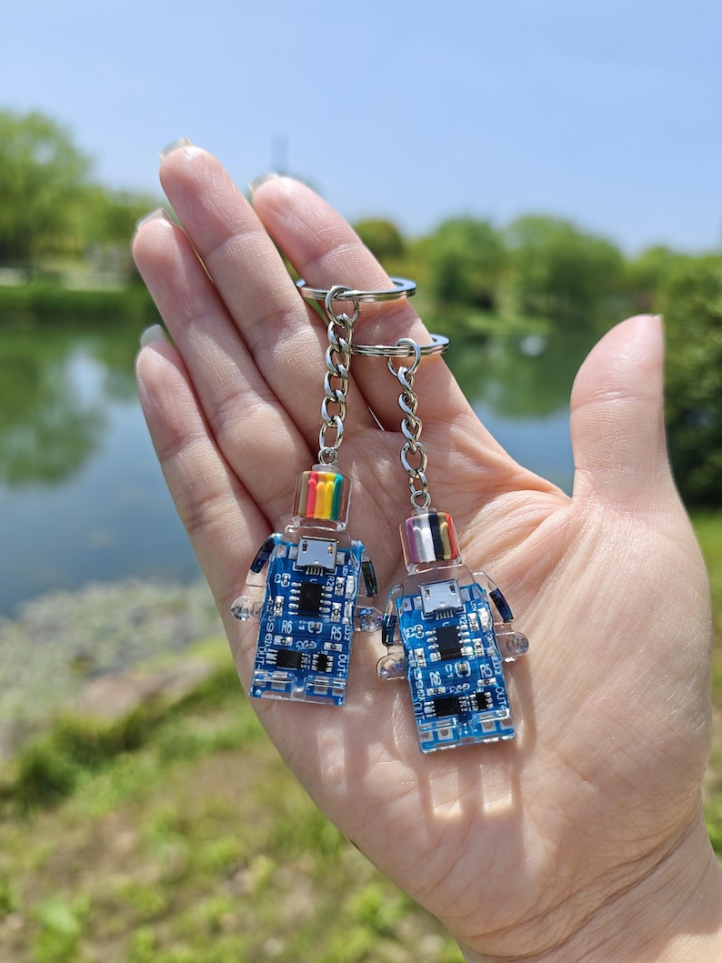 May include: Two keychains featuring a robot design with a blue circuit board body and clear plastic limbs. Each robot has a rainbow or clear head and is attached to a silver chain and keyring. The background shows a lake and greenery.