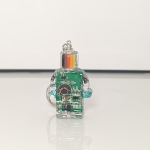 May include: A clear, miniature figurine in the shape of a person, featuring a green circuit board design inside. The head has a silver keychain attachment and a stripe of black, red, yellow, and gold. The figurine has a blue accent on the side.
