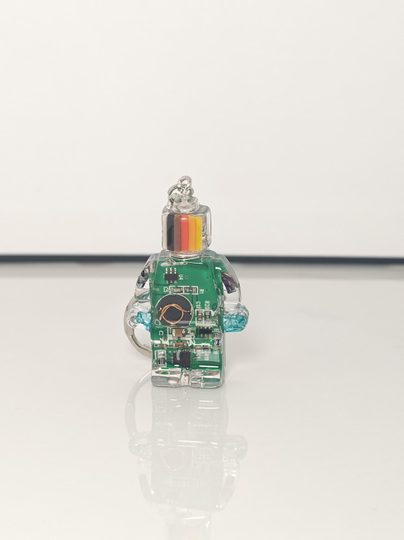 May include: A clear, miniature figurine in the shape of a person, featuring a green circuit board design inside. The head has a silver keychain attachment and a stripe of black, red, yellow, and gold. The figurine has a blue accent on the side.
