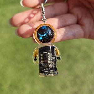 May include: An astronaut-shaped keychain made of clear resin containing a black circuit board. The helmet features a dark blue and black space-themed graphic with stars. The figure has gold-colored wire coils at the shoulders and a metal ring.