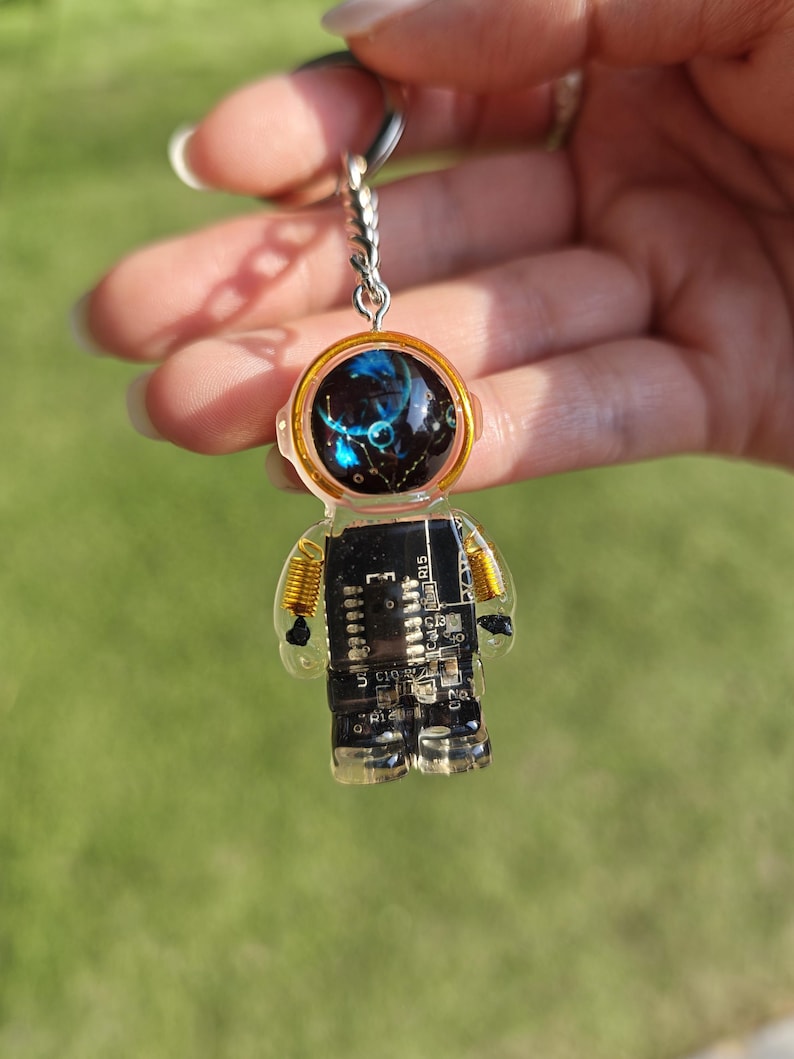May include: An astronaut-shaped keychain made of clear resin containing a black circuit board. The helmet features a dark blue and black space-themed graphic with stars. The figure has gold-colored wire coils at the shoulders and a metal ring.