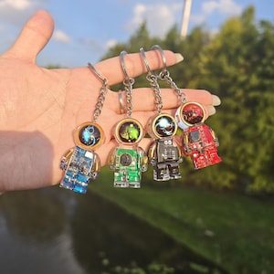 May include: Four astronaut keychains featuring transparent bodies with visible internal circuitry and colorful helmets. The figures are blue, green, black, and red, each attached to a silver metal chain and ring for secure attachment to keys.