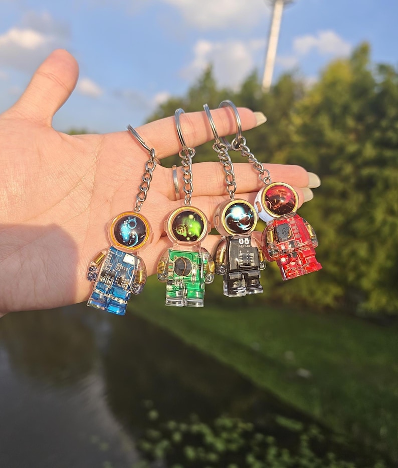 May include: Four astronaut keychains featuring transparent bodies with visible internal circuitry and colorful helmets. The figures are blue, green, black, and red, each attached to a silver metal chain and ring for secure attachment to keys.
