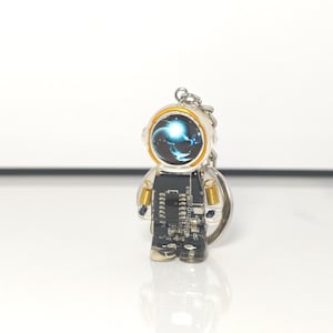 May include: A keychain shaped like an astronaut figure with a black circuit board body, gold-colored shoulder coils, and a helmet visor displaying a blue and white swirling nebula graphic. The figure is made of clear resin and includes a metal ring.