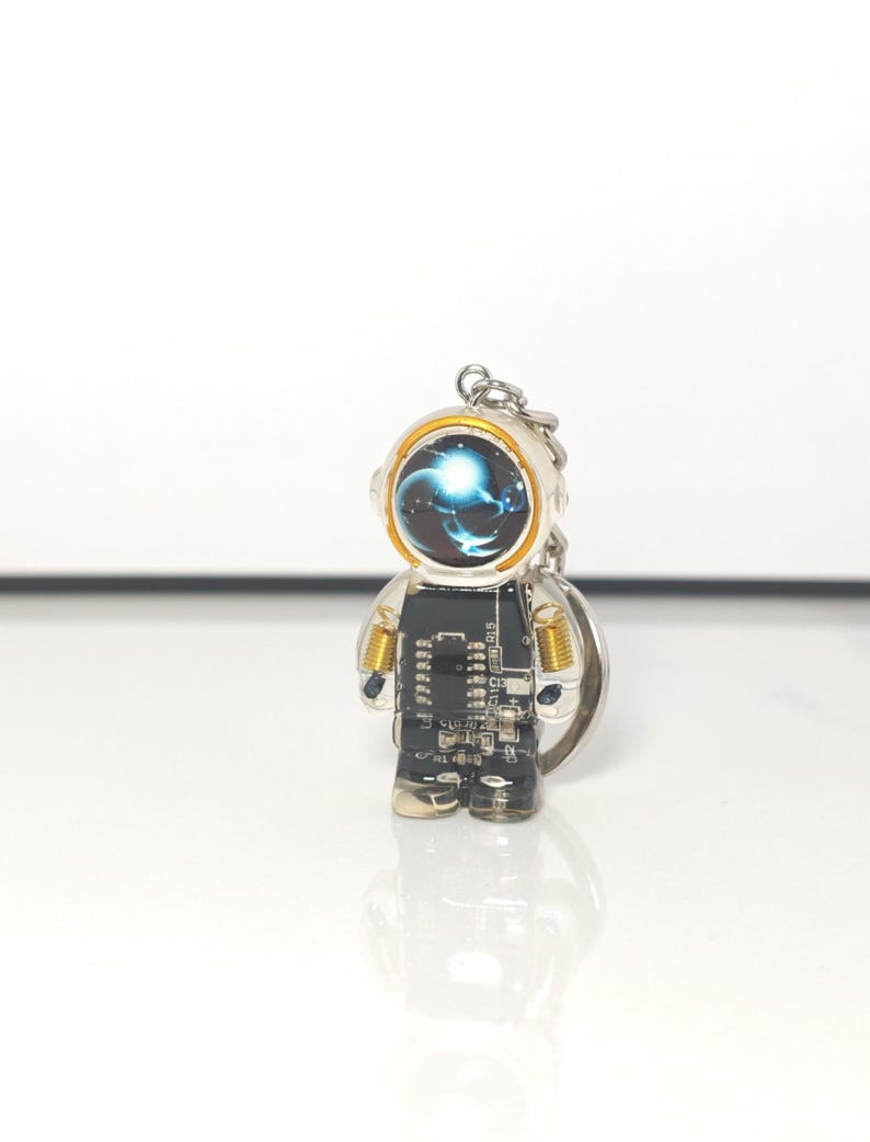 May include: A keychain shaped like an astronaut figure with a black circuit board body, gold-colored shoulder coils, and a helmet visor displaying a blue and white swirling nebula graphic. The figure is made of clear resin and includes a metal ring.