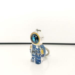 May include: A keychain featuring a small, translucent blue astronaut figure. The astronaut has a white helmet with a dark visor displaying a blue nebula pattern, a blue suit with white circuit board detailing, and a small gold-colored spring accessory.