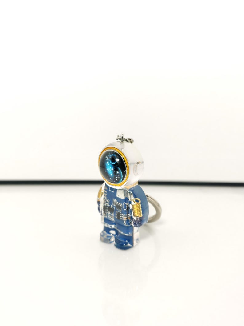 May include: A keychain featuring a small, translucent blue astronaut figure. The astronaut has a white helmet with a dark visor displaying a blue nebula pattern, a blue suit with white circuit board detailing, and a small gold-colored spring accessory.