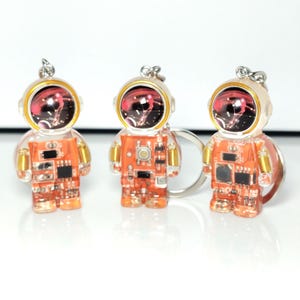 May include: Three astronaut-shaped keychains featuring translucent orange bodies with visible internal electronic circuit board components, gold-colored coil details on the shoulders, and a dark helmet visor reflecting a red and white nebula pattern.