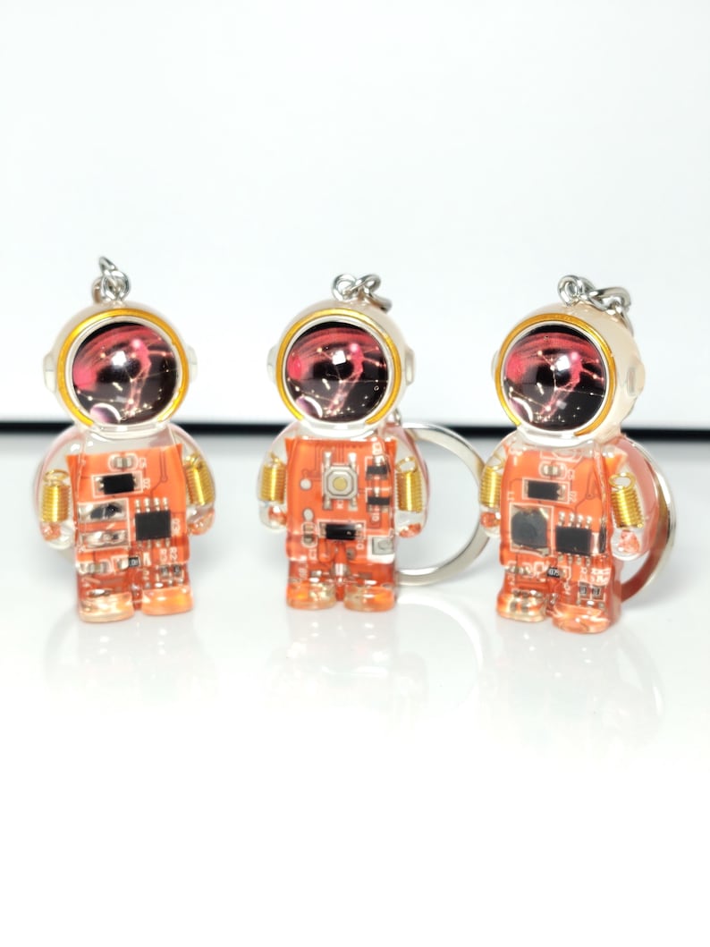 May include: Three astronaut-shaped keychains featuring translucent orange bodies with visible internal electronic circuit board components, gold-colored coil details on the shoulders, and a dark helmet visor reflecting a red and white nebula pattern.