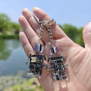 May include: Two clear acrylic keychains in the shape of a person, each with a circuit board design. The keychains are attached to a silver chain and ring. The tops of the keychains have a colorful, striped design.