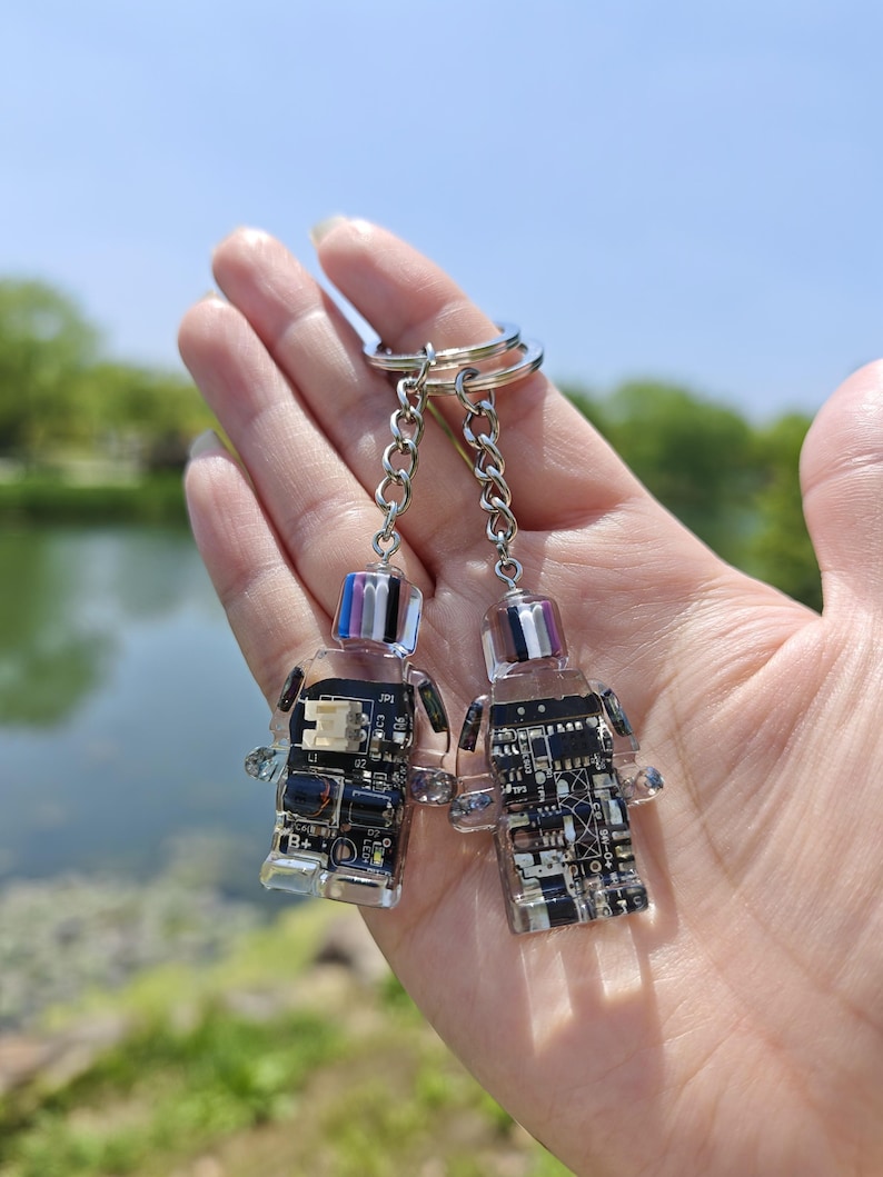 May include: Two clear acrylic keychains in the shape of a person, each with a circuit board design. The keychains are attached to a silver chain and ring. The tops of the keychains have a colorful, striped design.