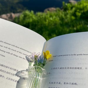 May include: A clear glass vase with a ruffled rim, holding delicate glass flowers in shades of pink, blue, and yellow. The vase sits on the open pages of a book, with text visible.