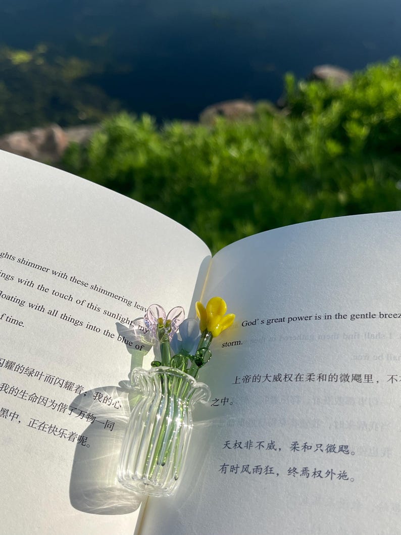 May include: A clear glass vase with a ruffled rim, holding delicate glass flowers in shades of pink, blue, and yellow. The vase sits on the open pages of a book, with text visible.