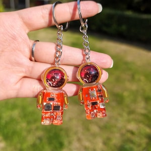 May include: Two astronaut-shaped keychains made of clear resin with embedded orange circuit boards. Each figure features a round helmet with a dark red, reflective visor and a metallic silver chain with a circular split ring for attachment.