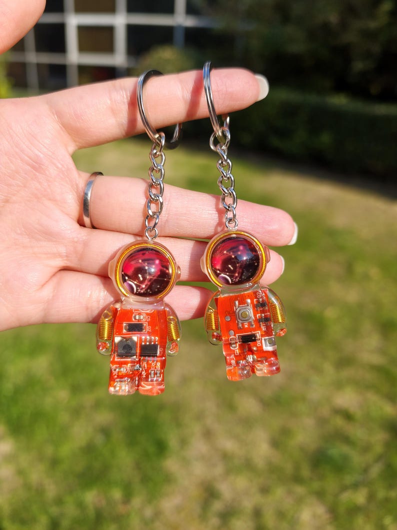 May include: Two astronaut-shaped keychains made of clear resin with embedded orange circuit boards. Each figure features a round helmet with a dark red, reflective visor and a metallic silver chain with a circular split ring for attachment.