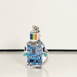 May include: A clear, robot-shaped keychain with a blue circuit board design inside. The robot's head features a rainbow stripe, and it has small, blue, crystal-like hands. A silver keyring is attached at the top.