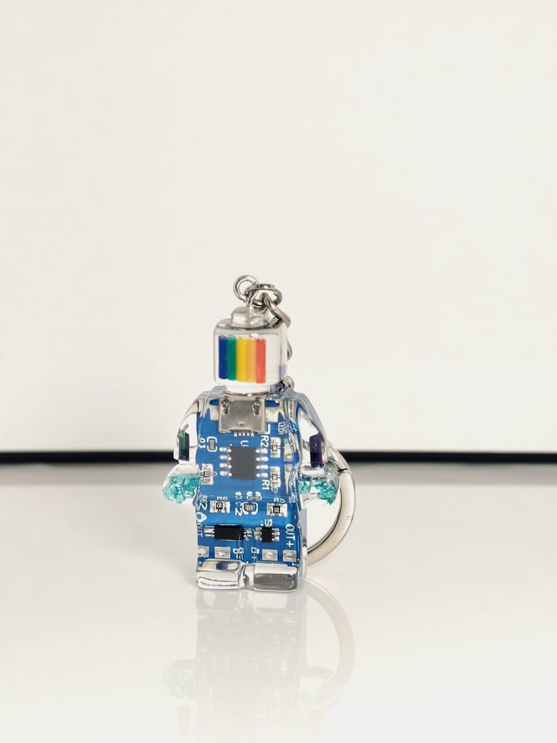 May include: A clear, robot-shaped keychain with a blue circuit board design inside. The robot's head features a rainbow stripe, and it has small, blue, crystal-like hands. A silver keyring is attached at the top.