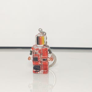May include: A clear, rectangular keychain in the shape of a person. The keychain has a red and black circuit board design inside. The top of the keychain has black, yellow, and red stripes. A silver keyring is attached.