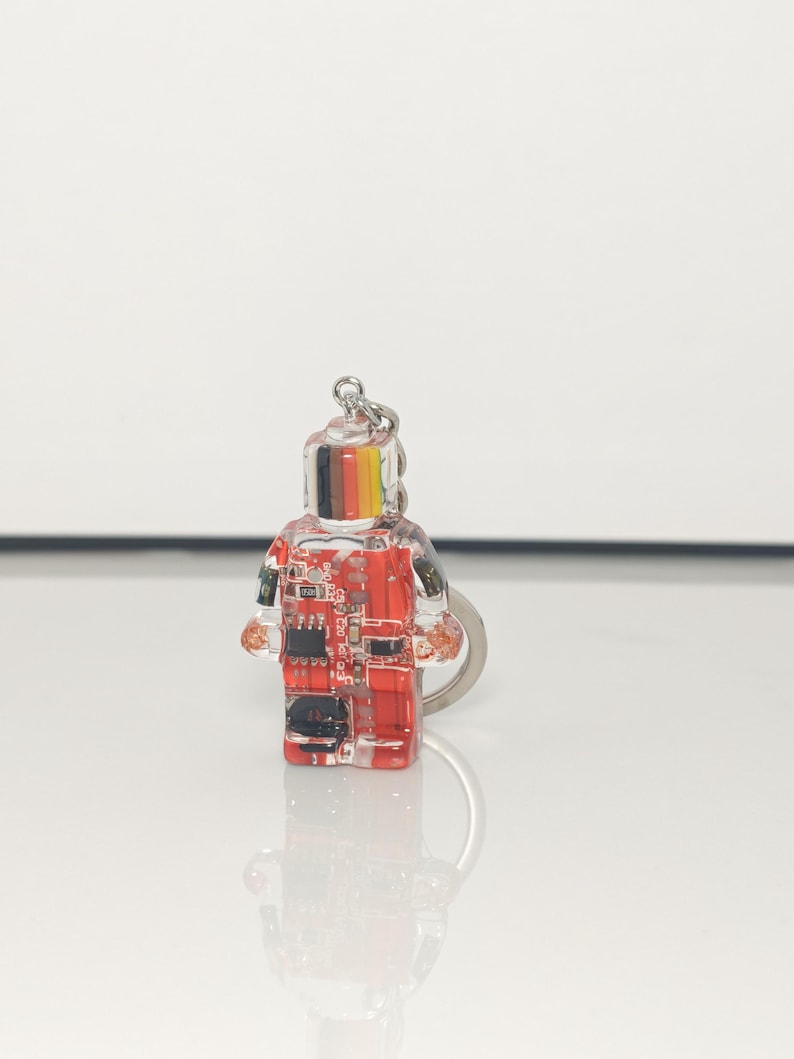 May include: A clear, rectangular keychain in the shape of a person. The keychain has a red and black circuit board design inside. The top of the keychain has black, yellow, and red stripes. A silver keyring is attached.