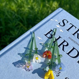 May include: Close-up of several glass flower pendants with green stems and colorful blossoms. The flowers are in shades of purple, white, red, yellow, and blue. The pendants are resting on a blue book with the title "ST BIRDS".