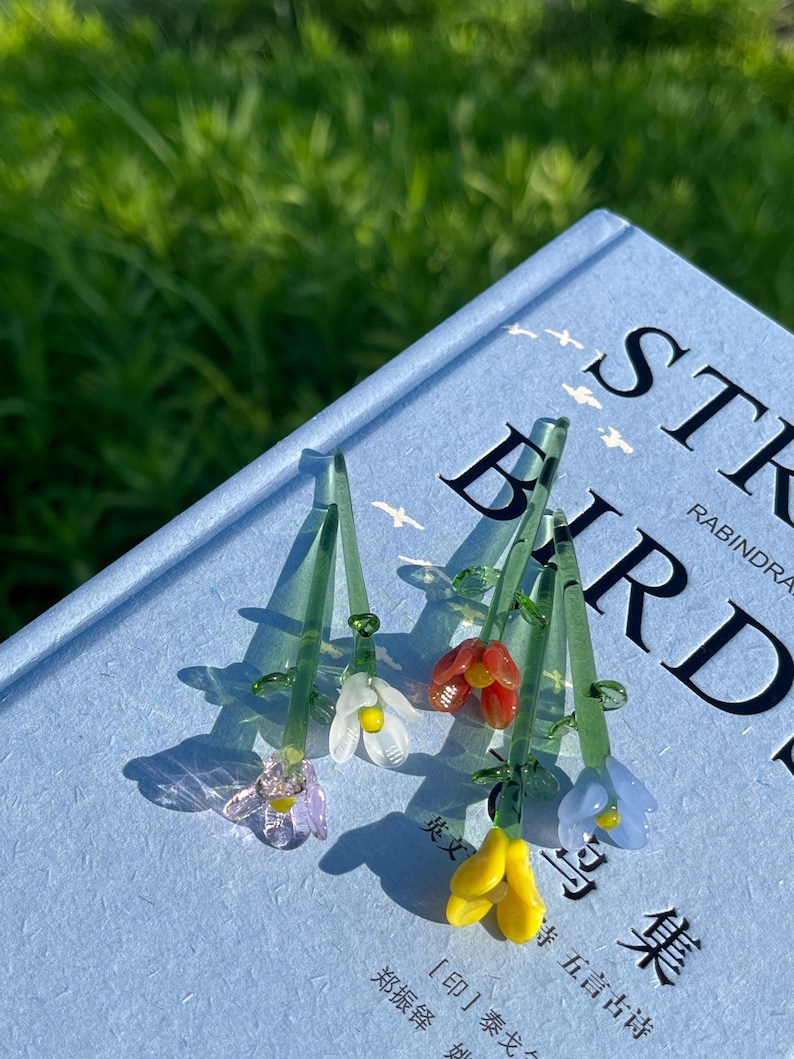 May include: Close-up of several glass flower pendants with green stems and colorful blossoms. The flowers are in shades of purple, white, red, yellow, and blue. The pendants are resting on a blue book with the title "ST BIRDS".