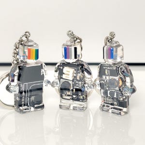 May include: Three clear robot-shaped keychains with silver chains. The robots have black bodies with circuit board designs and colorful striped heads. The keychains are displayed on a white surface.