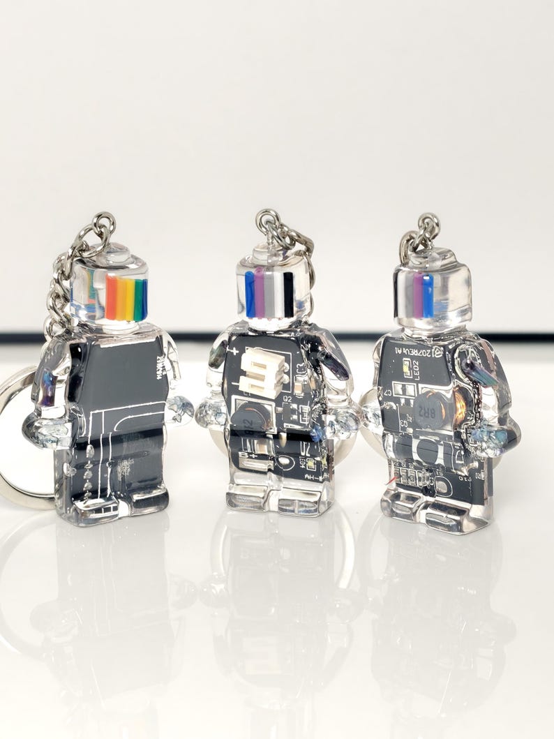 May include: Three clear robot-shaped keychains with silver chains. The robots have black bodies with circuit board designs and colorful striped heads. The keychains are displayed on a white surface.