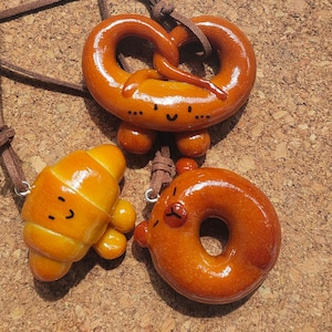 Pode incluir: Três pingentes castanhos com tema de comida em cordões castanhos. Os pingentes incluem um pretzel, um croissant e um donut, cada um com um rosto sorridente. O pretzel mede aproximadamente 5 cm de largura.