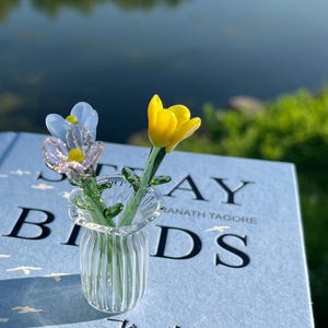 May include: A small glass vase with two miniature glass flowers, one yellow and one blue, on a book. The book has the title "STRAY BIRDS" and text in English and another language. The vase is clear.