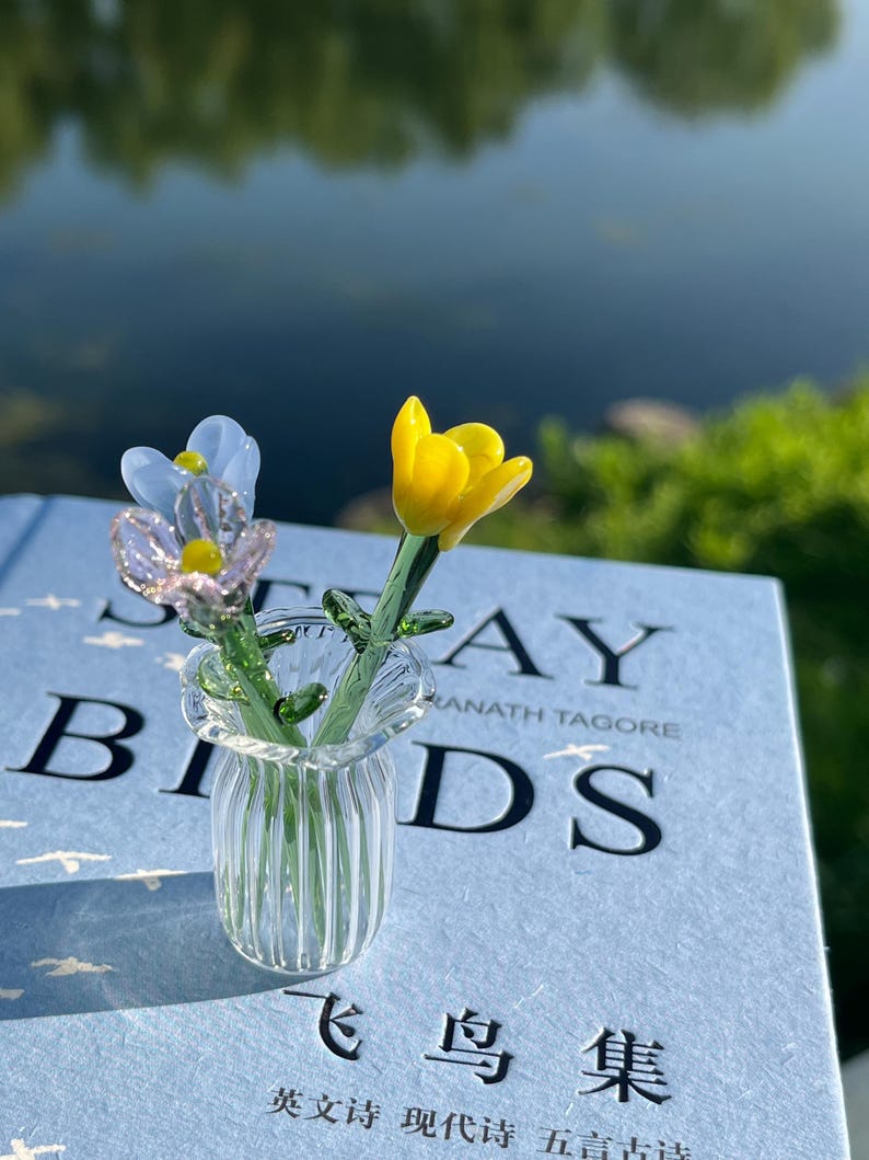 May include: A small glass vase with two miniature glass flowers, one yellow and one blue, on a book. The book has the title "STRAY BIRDS" and text in English and another language. The vase is clear.