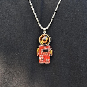 May include: A silver-toned box chain necklace featuring a pendant shaped like an astronaut. The pendant is constructed from a red circuit board with gold-colored coil accents on the arms and a clear, rounded helmet dome revealing a dark, detailed interior.