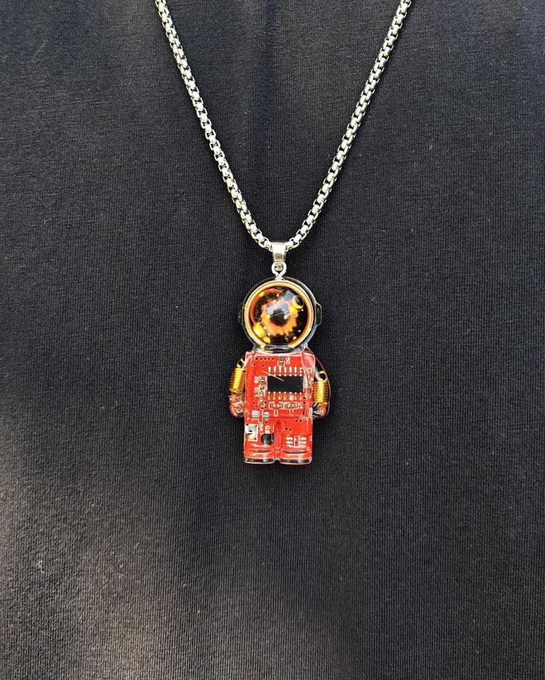 May include: A silver-toned box chain necklace featuring a pendant shaped like an astronaut. The pendant is constructed from a red circuit board with gold-colored coil accents on the arms and a clear, rounded helmet dome revealing a dark, detailed interior.