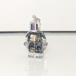 May include: A clear, robot-shaped keychain with a silver keyring. The robot's body is made of clear resin, showcasing internal electronic components. The head features vertical stripes of blue, pink, and white. The keychain is on a white surface.