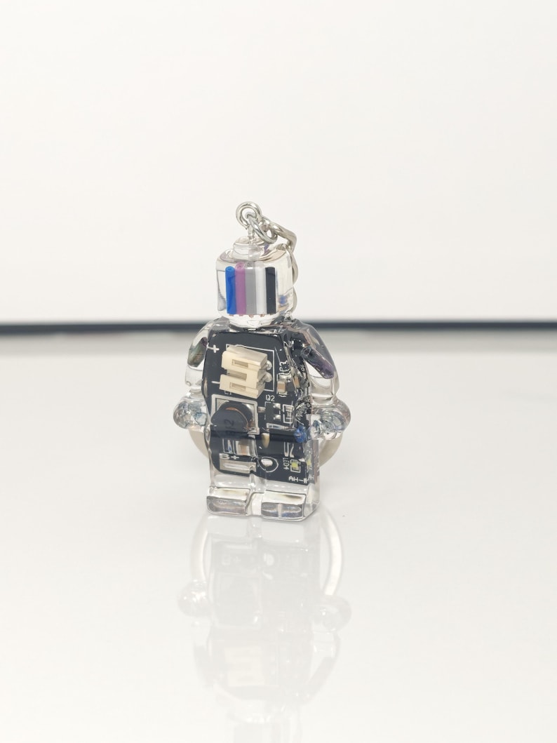May include: A clear, robot-shaped keychain with a silver keyring. The robot's body is made of clear resin, showcasing internal electronic components. The head features vertical stripes of blue, pink, and white. The keychain is on a white surface.