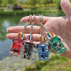 May include: Four colorful robot-shaped keychains. Each keychain features a clear body with internal circuit board details and a rainbow-colored top. The keychains are red, black, blue, and green, attached to silver keyrings.